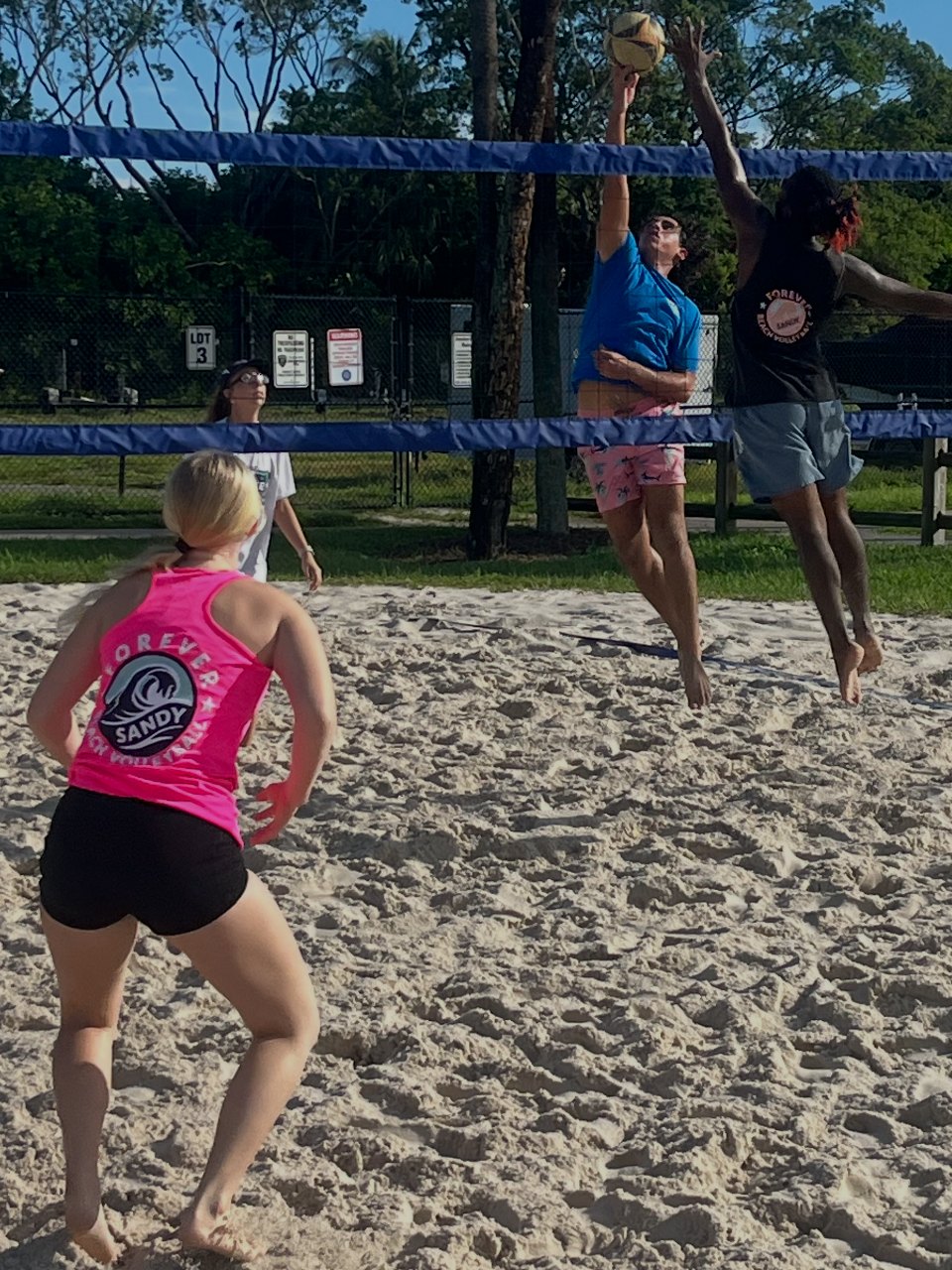 A Forever Sandy player in a pink team tank watching a teammate swing at the net while a blocker goes up to stop it.
